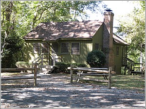A small green cottage with a chimney, surrounded by trees and a wooden fence, featuring a front porch and picnic tables.