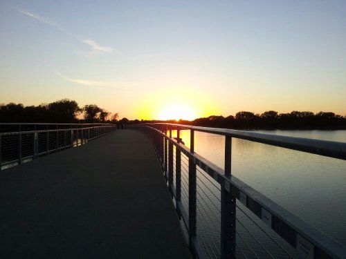 A serene walkway along a river at sunset, with trees silhouetted against the colorful sky.