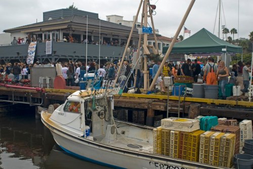 A fishing boat docked near a busy waterfront restaurant with people gathered outside on a cloudy day.