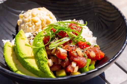 A black bowl filled with poke, avocado slices, green onions, and rice, garnished with sesame seeds and diced tomatoes.