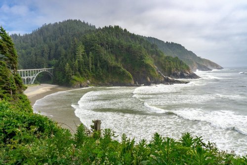 A scenic coastal view featuring a bridge, lush green hills, and waves crashing on a sandy beach.