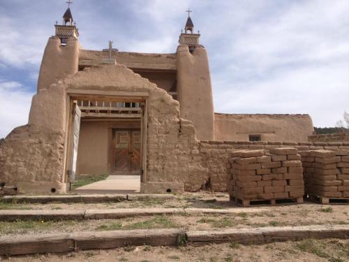 Adobe building with a wooden door, cross-topped towers, and stacked bricks in front, set against a cloudy sky.