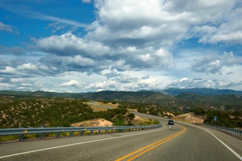 A winding road through green hills under a blue sky with fluffy clouds. A car drives along the scenic route.