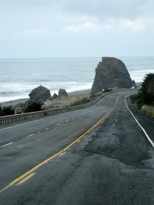 A coastal road curves along the shore, with rocky formations and the ocean in the background under a cloudy sky.