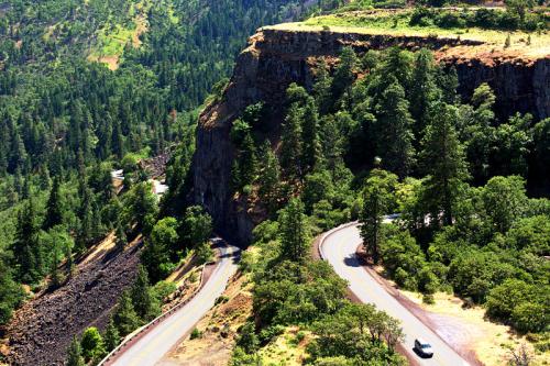 A winding road curves through lush green trees and rocky cliffs in a scenic mountainous landscape.