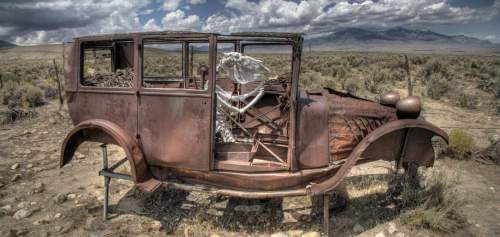 An old, rusted car sits abandoned in a desert landscape under a cloudy sky.