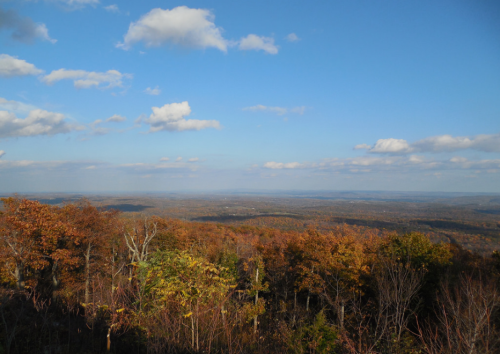 A panoramic view of autumn foliage with rolling hills under a blue sky and scattered clouds.
