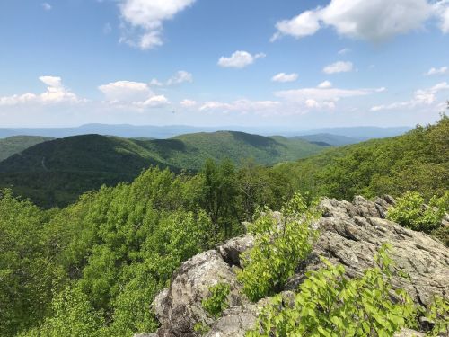 A scenic view of rolling green mountains under a blue sky with fluffy clouds, seen from a rocky outcrop.