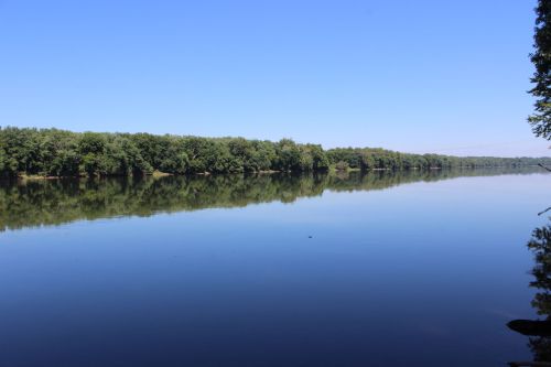 A calm river reflects a clear blue sky and lush green trees along its banks.