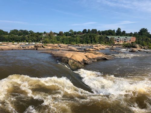 A river flows over rocky formations, with greenery and buildings visible in the background under a clear blue sky.