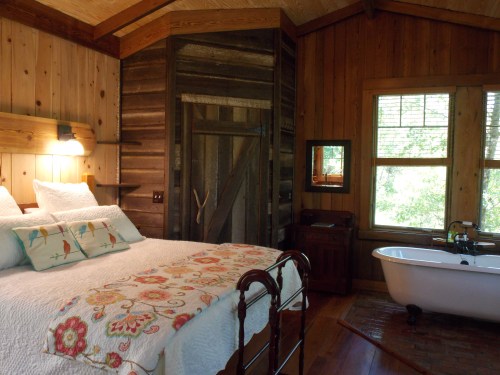 Cozy cabin interior featuring a bed with colorful bedding, a vintage bathtub, and wooden walls. Natural light from windows.