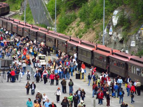 A crowded train station with a long train and many people waiting and walking around in a scenic outdoor setting.