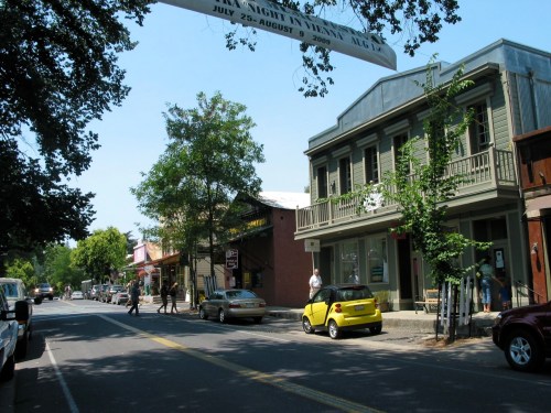 A sunny street scene with trees, historic buildings, and parked cars, featuring a yellow car and pedestrians.