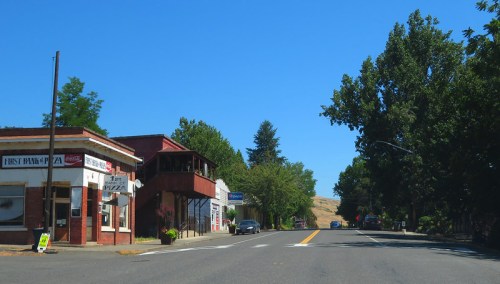 A quiet street scene featuring small shops, trees, and clear blue skies in a rural town.