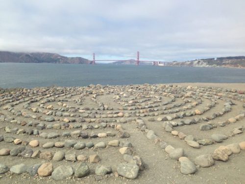 A stone labyrinth on a beach with the Golden Gate Bridge in the background and a cloudy sky above.