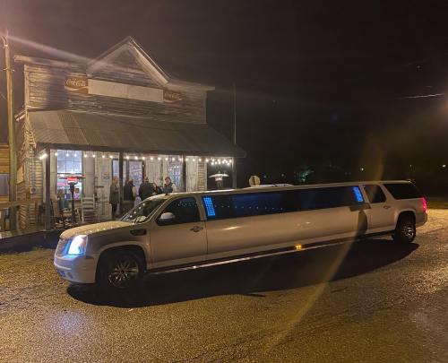 A white limousine parked outside a rustic building at night, with people gathered nearby.