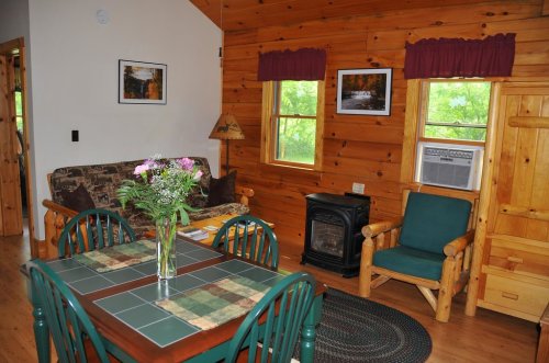Cozy cabin interior with wooden walls, a dining table, sofa, and a small heater, surrounded by windows and greenery.