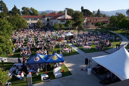 Aerial view of a large outdoor event with tables, tents, and attendees in a park-like setting. Mountains in the background.