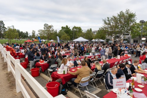 A large outdoor gathering with people seated at tables, enjoying food and festivities under cloudy skies.