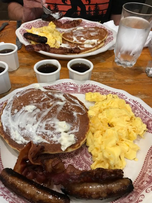 Two plates of breakfast featuring pancakes with butter, scrambled eggs, bacon, and sausage, with syrup on the side.
