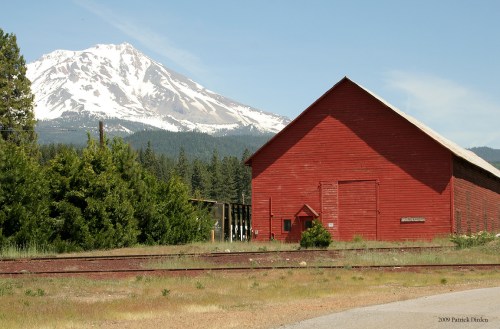 A red barn stands near a railway track, with a snow-capped mountain in the background and trees surrounding the area.
