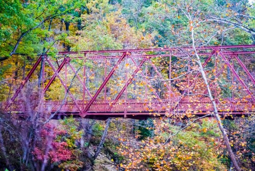 A red metal bridge spans a creek, surrounded by vibrant autumn foliage in various shades of orange, yellow, and green.