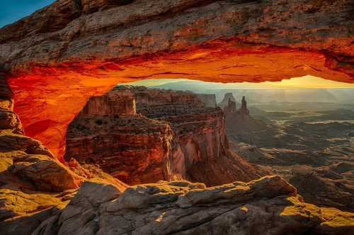 A stunning view through a rock arch, showcasing layered cliffs and a vibrant sunset over a vast canyon landscape.