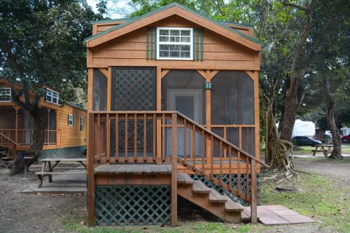 A wooden cabin with a screened porch and steps, surrounded by trees in a campsite setting.