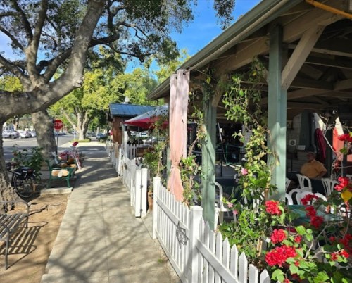 A sunny sidewalk lined with flowers, leading to a charming outdoor café with tables and umbrellas.