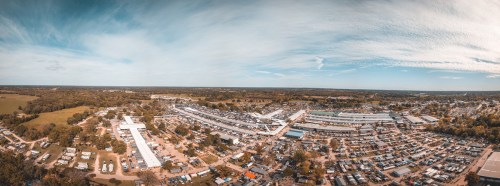 Aerial view of a large outdoor market with numerous stalls and vehicles, surrounded by green fields and blue sky.
