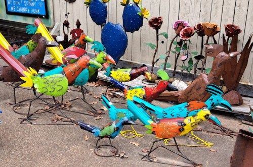 Colorful metal sculptures of birds and flowers arranged in a garden setting, showcasing vibrant artistic designs.