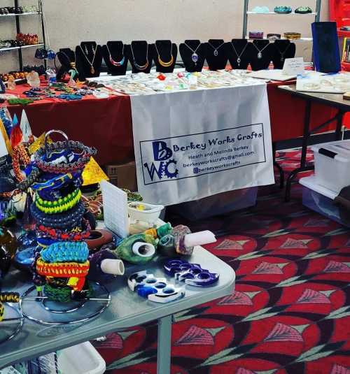 A craft fair display featuring jewelry on stands, colorful glass items, and a table with a banner for Berkey Works Crafts.