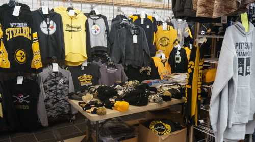 A display of Pittsburgh Steelers and baseball merchandise, including shirts, hoodies, and hats in black and gold.