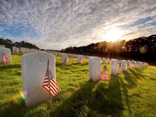 Graves marked with flags in a serene cemetery at sunset, with clouds and trees in the background.