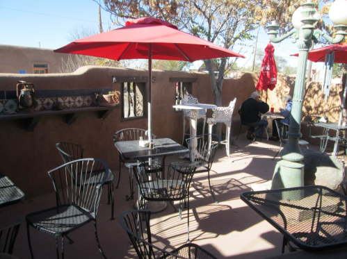 Outdoor patio with black metal tables and chairs, red umbrellas, and a warm, sunny atmosphere.