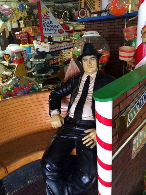 A life-sized mannequin in a suit and hat sits on a bench in a colorful candy shop filled with sweets and treats.