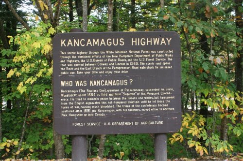 Sign about Kancamagus Highway in the White Mountains, detailing its history and connection to the region. Surrounded by trees.