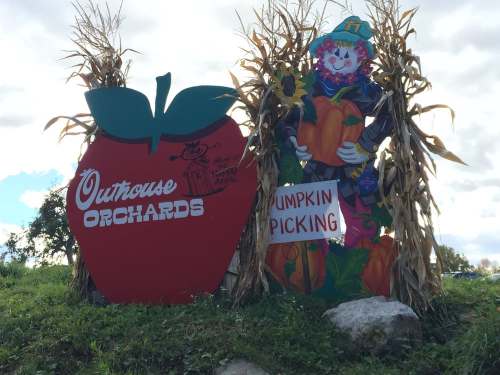 Sign for Outhouse Orchards featuring a large apple and a scarecrow holding pumpkins, surrounded by corn stalks.