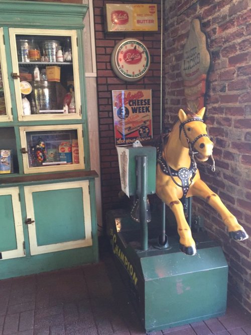 A vintage horse ride-on toy next to a green cabinet and retro advertisements on a brick wall.