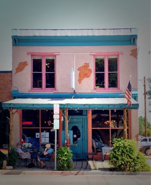 A colorful storefront with pink windows and a blue awning, featuring outdoor seating and a small American flag.