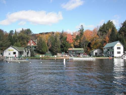 Lakeside view of buildings surrounded by colorful autumn trees and calm water under a blue sky.