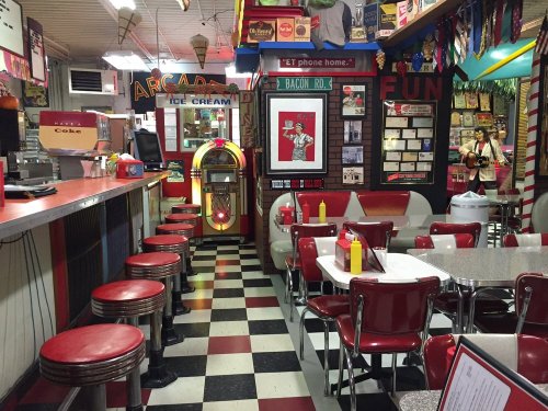 A retro diner interior with red and white decor, checkered floor, jukebox, and tables with condiments.