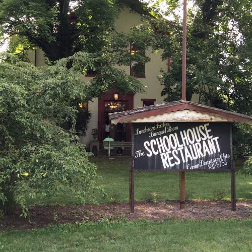 A sign for "The Schoolhouse Restaurant" in front of a green, leafy area and a building in the background.