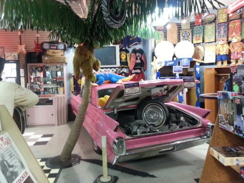 A pink vintage car with an open trunk, surrounded by quirky decor, including a palm tree and various collectibles.