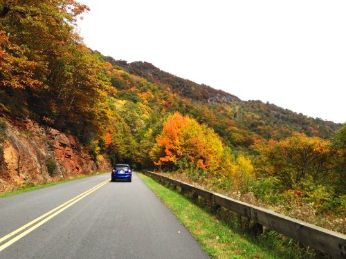 A blue car drives along a scenic road lined with vibrant autumn trees and mountains in the background.