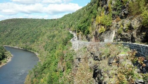 A winding road along a rocky cliffside, overlooking a river and lush green trees under a partly cloudy sky.