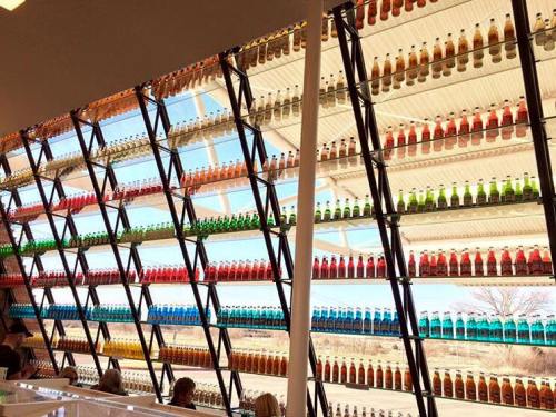 A colorful display of soda bottles arranged in a modern, angular shelving unit against a bright window.