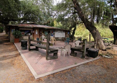 A rustic outdoor gathering space with benches and chairs under trees, featuring a small building in the background.