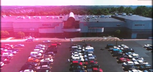 Aerial view of a large shopping center with a busy parking lot filled with cars on a sunny day.