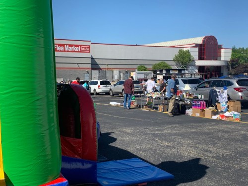 A bustling flea market scene with vendors and shoppers, featuring a colorful bounce house in the foreground.